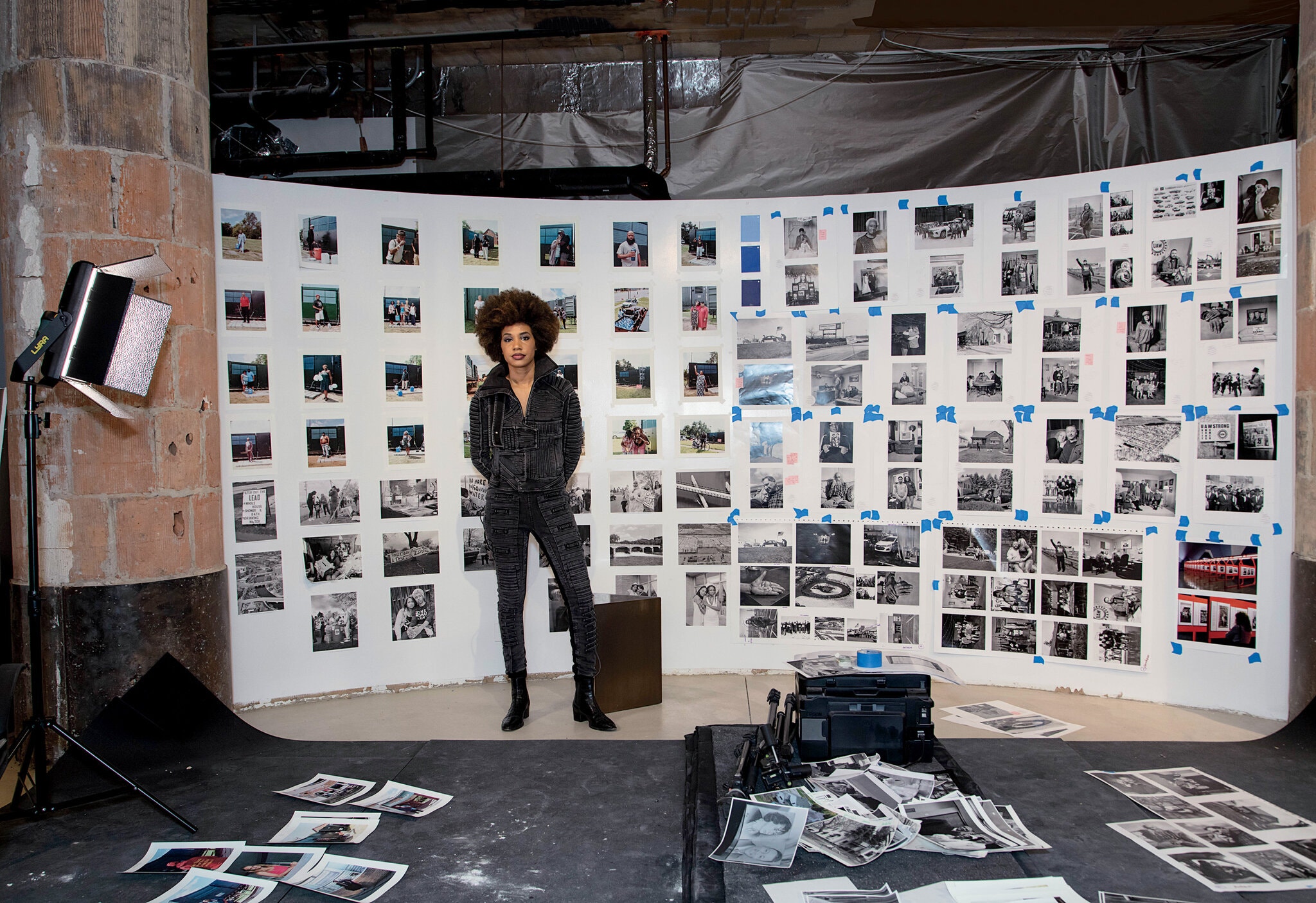 In an industrial studio space, a Black woman with an afro, wearing a black jean jacket and pants stands in front of a white wall filled with columns of photographs taped up with blue tape. Around her, other photoraphs are arranged on the floor. A large light stands to the left.