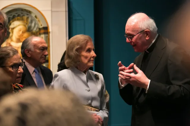 A group of light-skinned older people stand close in an art gallery listening as a Monsignor speaks to them. In the background is an artwork depicting Mary and Jesus; the wall next to it is painted teal.