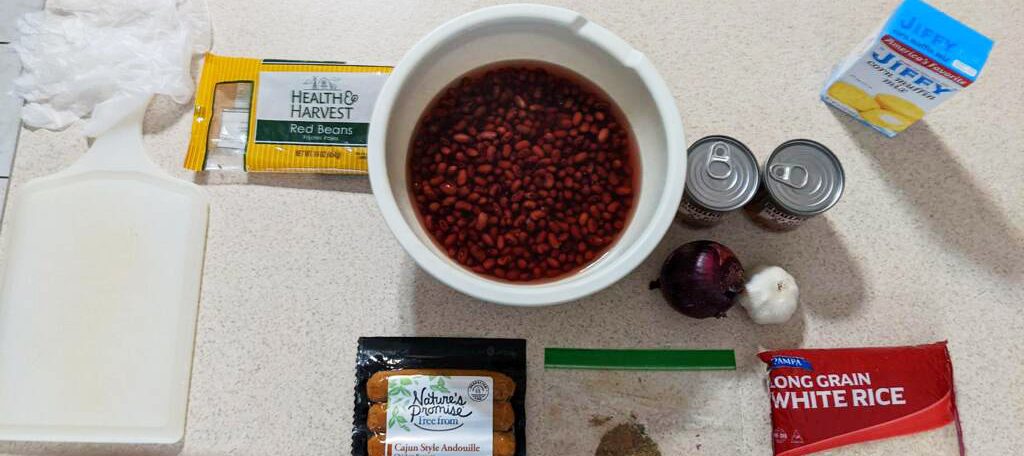 An aerial view of a white countertop with a bowl of red beans and other ingredients laid out on it, including a couple of cans, onion, garlic, a bag of rice, a box of corn muffin mix, and sausages.