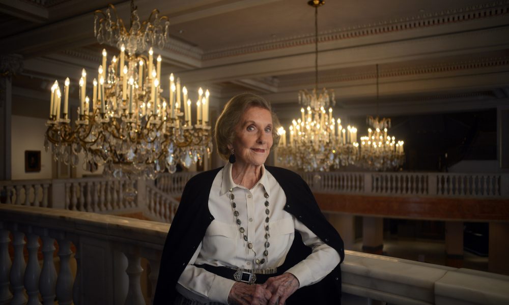 Wilhelmina Cole Holladay leans against a railing with a slight smile. She is a light-skinned, older woman with short, gray hair, and she wears a collared white shirt and black cardigan. Ornate chandeliers can be seen behind her.