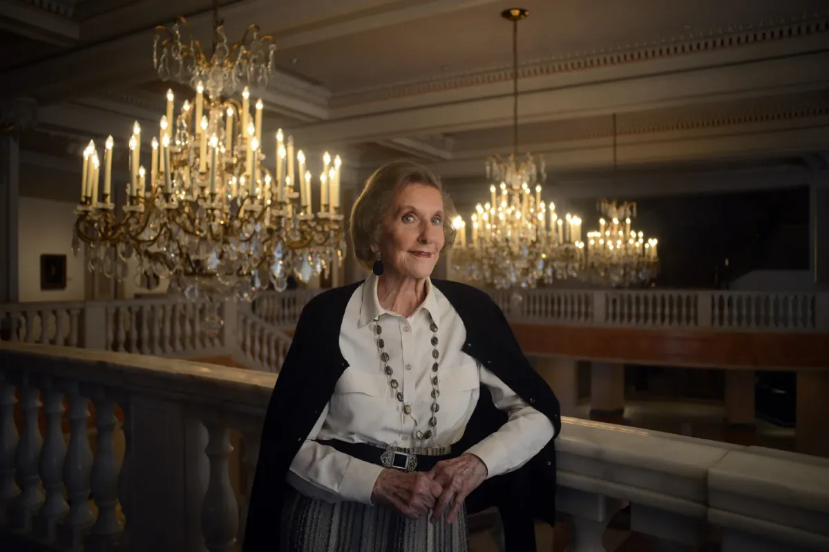 Wilhelmina Cole Holladay leans against a railing with a slight smile. She is a light-skinned, older woman with short, gray hair, and she wears a collared white shirt and black cardigan. Ornate chandeliers can be seen behind her.