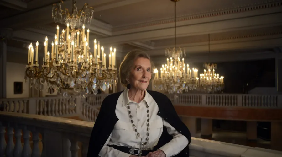 Wilhelmina Cole Holladay leans against a railing with a slight smile. She is a light-skinned, older woman with short, gray hair, and she wears a collared white shirt and black cardigan. Ornate chandeliers can be seen behind her.