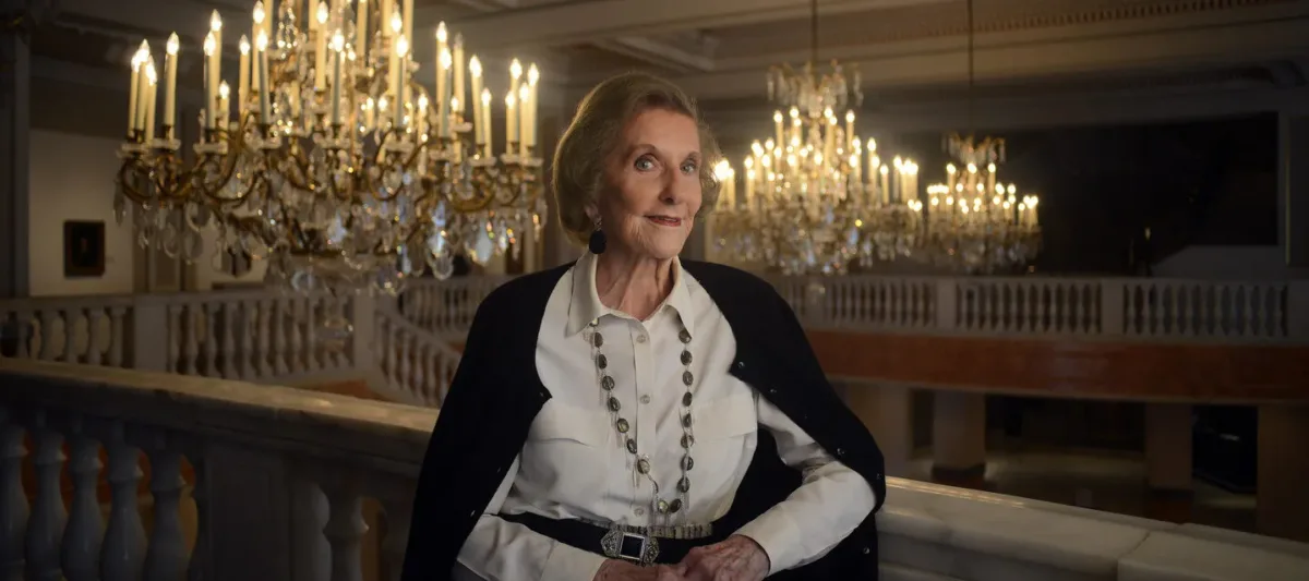 Wilhelmina Cole Holladay leans against a railing with a slight smile. She is a light-skinned, older woman with short, gray hair, and she wears a collared white shirt and black cardigan. Ornate chandeliers can be seen behind her.