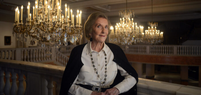 Wilhelmina Cole Holladay leans against a railing with a slight smile. She is a light-skinned, older woman with short, gray hair, and she wears a collared white shirt and black cardigan. Ornate chandeliers can be seen behind her.