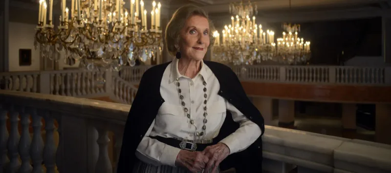 Wilhelmina Cole Holladay leans against a railing with a slight smile. She is a light-skinned, older woman with short, gray hair, and she wears a collared white shirt and black cardigan. Ornate chandeliers can be seen behind her.