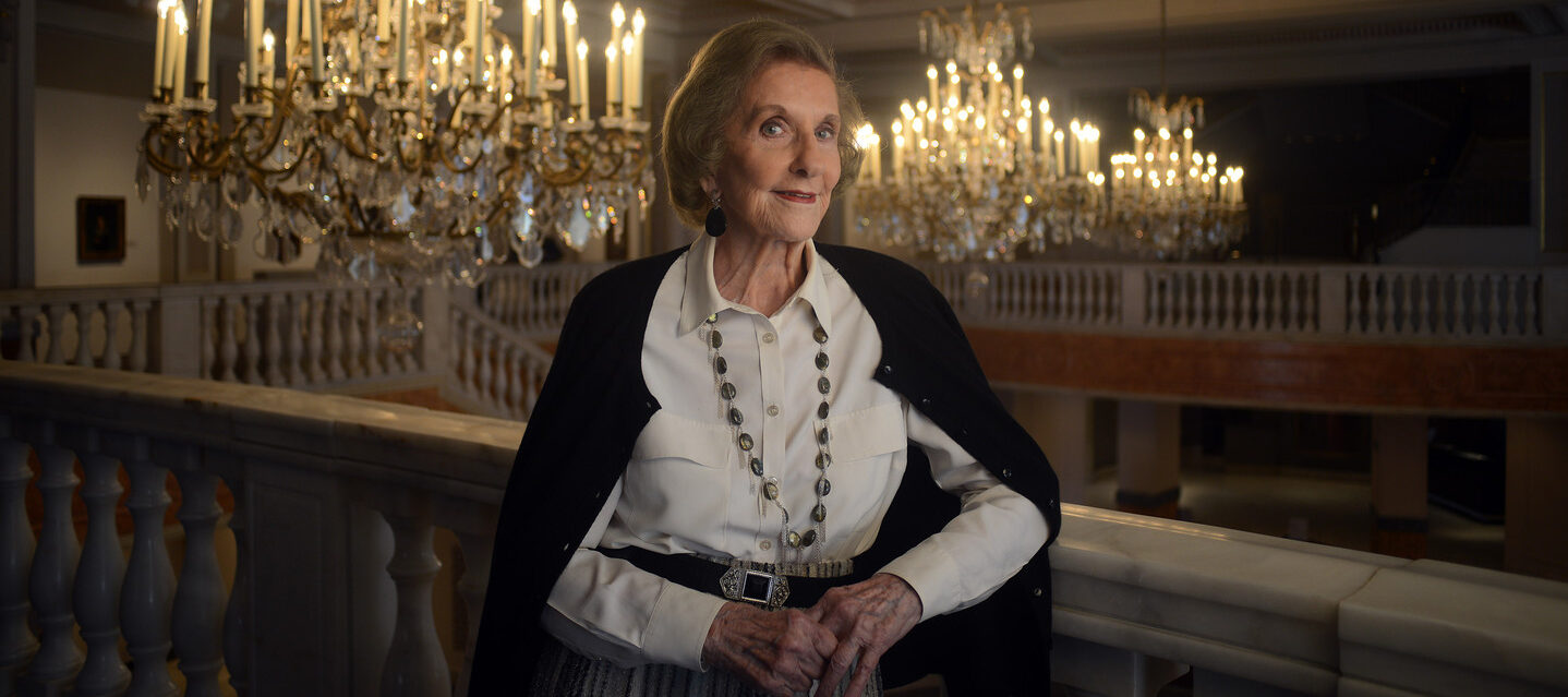 Wilhelmina Cole Holladay leans against a railing with a slight smile. She is a light-skinned, older woman with short, gray hair, and she wears a collared white shirt and black cardigan. Ornate chandeliers can be seen behind her.