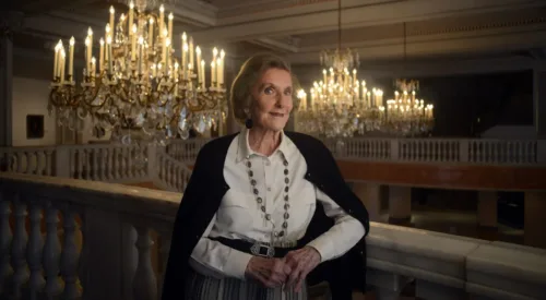 Wilhelmina Cole Holladay leans against a railing with a slight smile. She is a light-skinned, older woman with short, gray hair, and she wears a collared white shirt and black cardigan. Ornate chandeliers can be seen behind her.