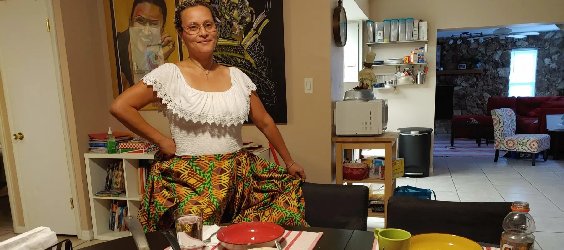 A medium skin-toned adult woman wearing traditional Puerto Rican dress—a white blouse with lace neck and brightly patterned skirt—stands smiling in front of her dining table in her home. The table is set with bowls and pots of ground beef, salsa, sauce, guacamole, and salad.