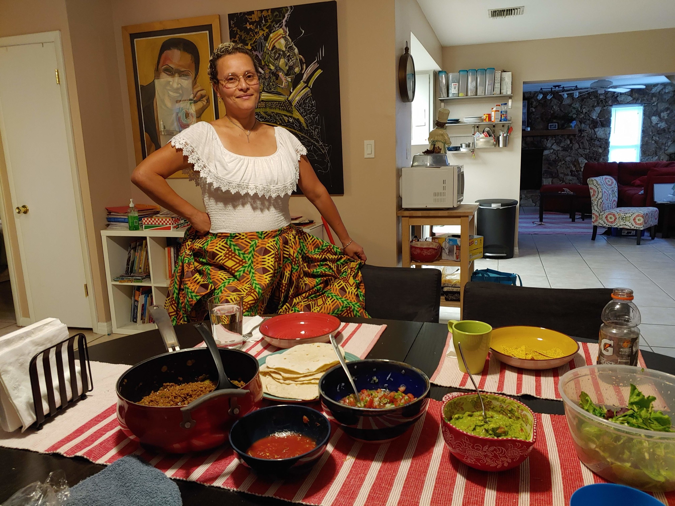 A medium skin-toned adult woman wearing traditional Puerto Rican dress—a white blouse with lace neck and brightly patterned skirt—stands smiling in front of her dining table in her home. The table is set with bowls and pots of ground beef, salsa, sauce, guacamole, and salad.