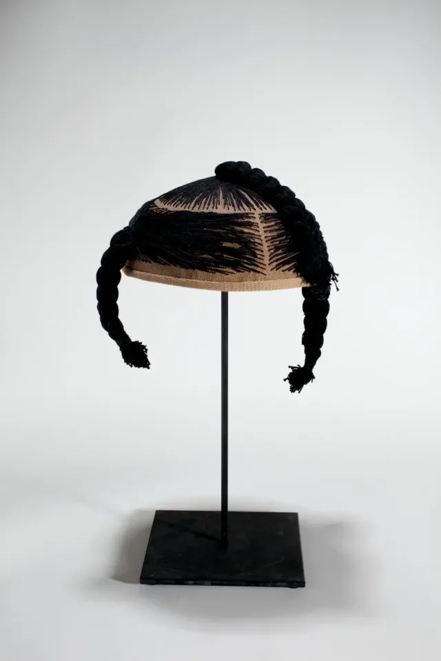 A wig cap sits mounted on a black stand. Black thread is stitched into the cap, mimicking hair. The thread forms three braids.