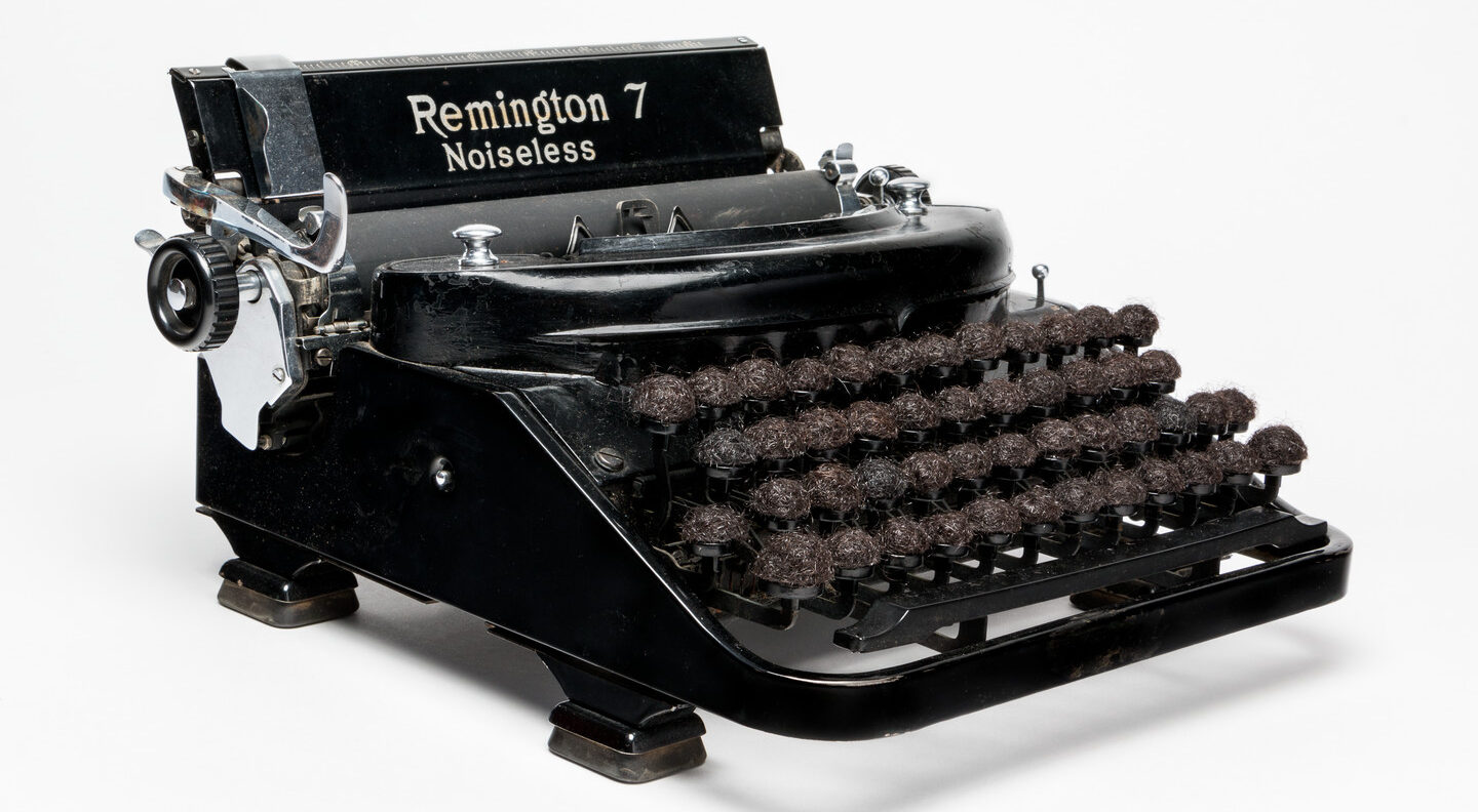 An antique, black and silver typewriter with the words 'Remington 7 Noiseless' emblazoned on the top. The lettered keys of the typewriter have been replaced with small balls of dark brown hair.