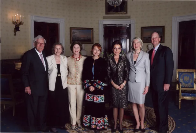 A formal photograph of seven light-skinned individuals—four women in the center, two men on either end—in the U.S. Presidential office. The woman in the very middle wears a traditional Mexican dress, to her left another woman wears a cheetah-patterned skirt and blazer set. The others are in various business dress. Everyone smiles at the camera.