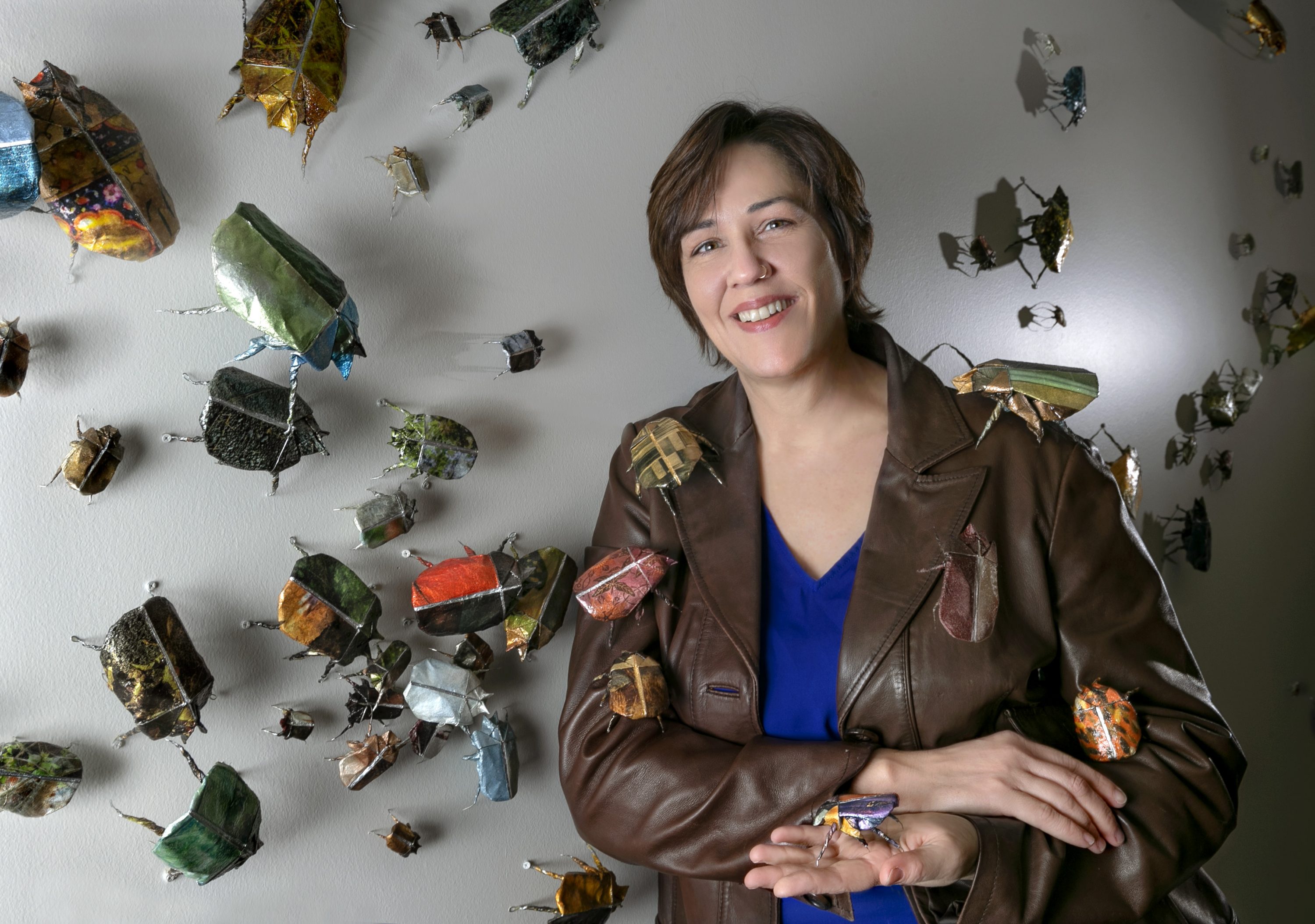 A light-skinned woman with short brown hair stands in front of a white wall to which paper beetles, sculpted out of photographs, are affixed. The woman holds a beetle in her open palm, while others are arranged atop her brown leather jacket. She smiles slightly at the camera while leaning against the wall.