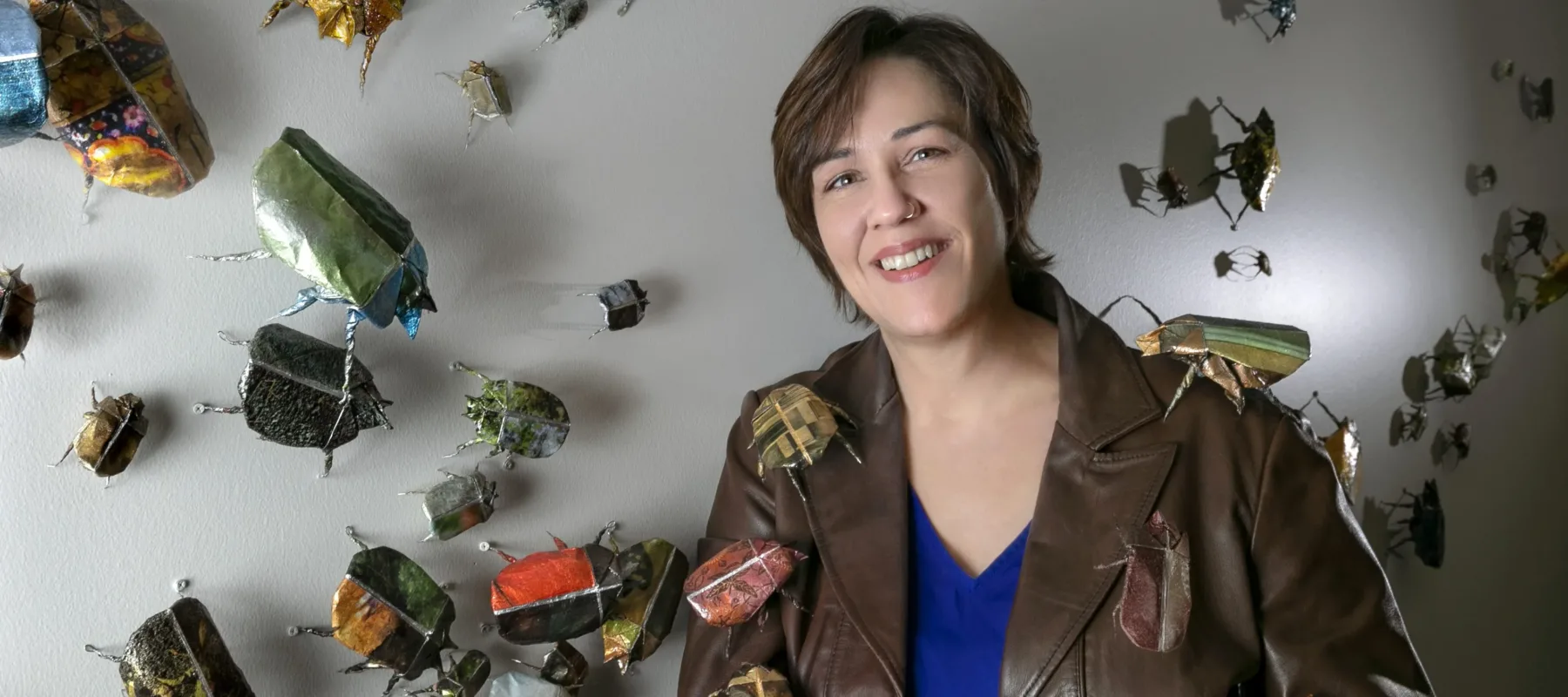 A light-skinned woman with short brown hair stands in front of a white wall to which paper beetles, sculpted out of photographs, are affixed. The woman holds a beetle in her open palm, while others are arranged atop her brown leather jacket. She smiles slightly at the camera while leaning against the wall.