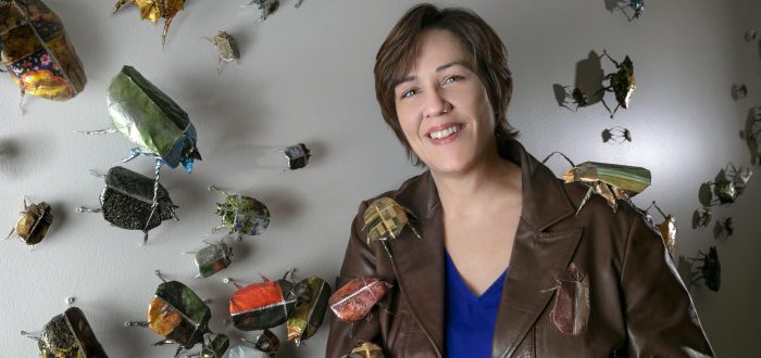 A light-skinned woman with short brown hair stands in front of a white wall to which paper beetles, sculpted out of photographs, are affixed. The woman holds a beetle in her open palm, while others are arranged atop her brown leather jacket. She smiles slightly at the camera while leaning against the wall.