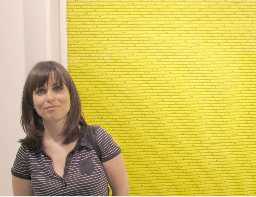 A medium-skinned woman stands in front of a bright yellow artwork. She has shoulder-length straight brown hair with bangs and wears a striped t-shirt. She smiles slightly.