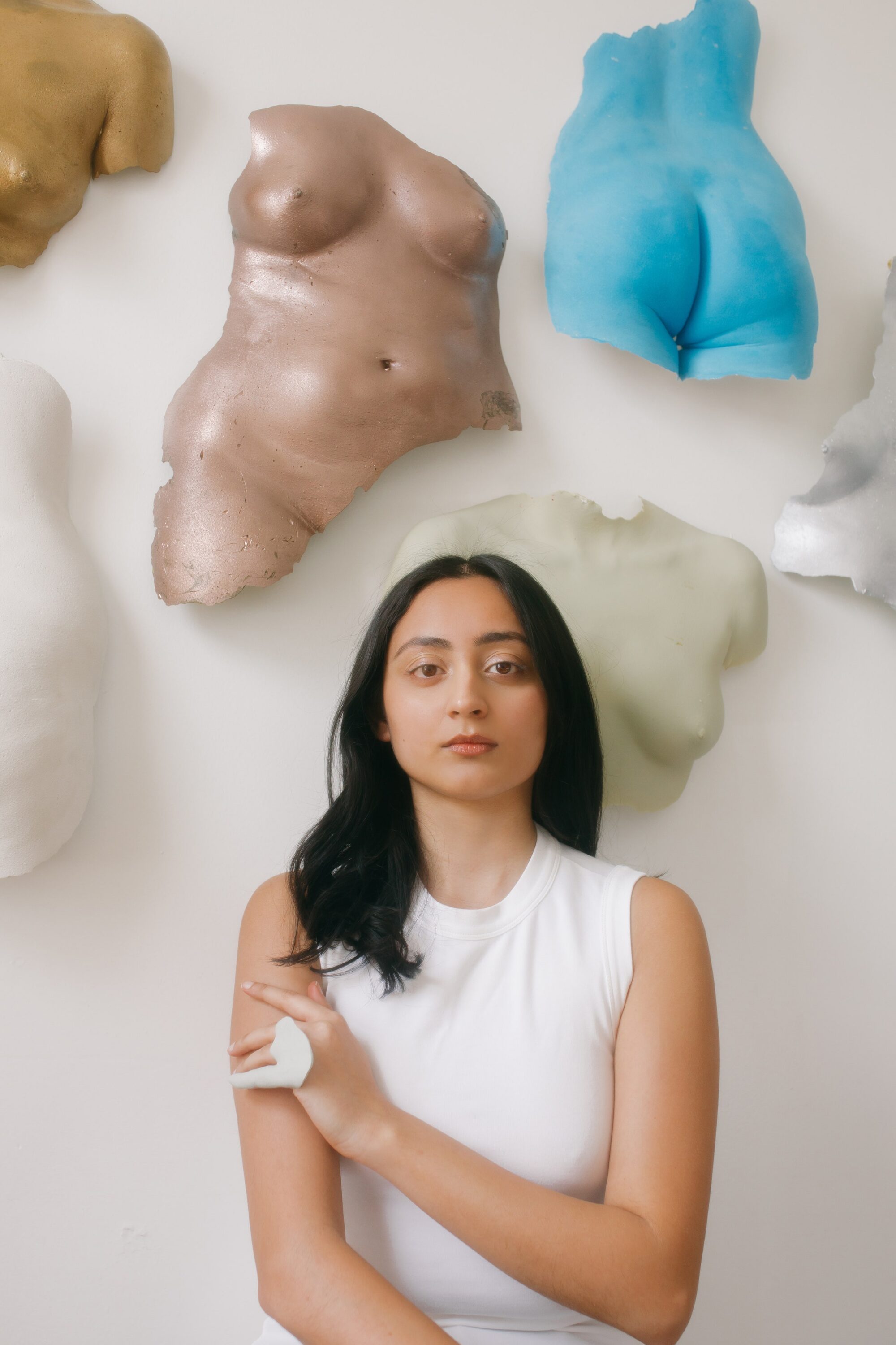 A medium-skinned woman wearing a white sleeveless shirt stands in front of a white wall. Above her, hung on the wall, are women's body casts in different colors: a blue lower back and butt, a bronze torso, a white chest.