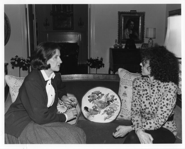 Two light-skinned women sit on a couch in a residence. The woman on the left wears a cardigan, blouse, and skirt and has chin-length straight brow hair. The woman on the right has curly shoulder-length hair, wears glasses, and a polka dotted top and black pants. They look at each other as they are in conversation. Four embroidered pillows sit on the couch behind and next to them.