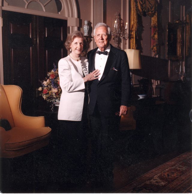 A light-skinned elderly couple formally pose for a photograph in an ornate room. They wear formal evening attire and smile subtly. The woman places her hand against the man's lower chest.