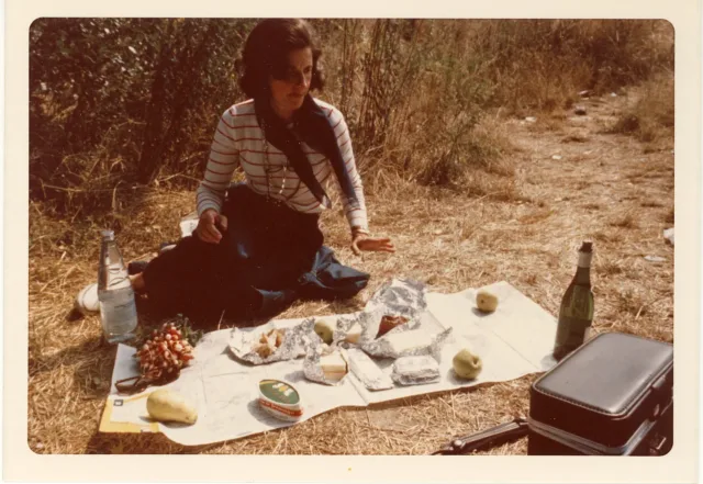 A vintage photograph of a light-skinned woman with short brown hair sitting on the ground with a map spread out in front of her. On top of the map is a picnic with apples, potatoes, radishes, sandwiches, a bottle of water, and a bottle of wine. There is a small black case in the bottom right corner of the photograph.