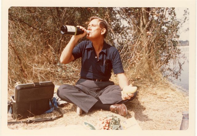 A vintage photograph of a light-skinned man with short light-colored hair sitting on the ground. The man drinks out of a full sized bottle of wine with a potato in his other hand. There is a small black case beside him and a bunch of radishes sitting on a map in front of him.