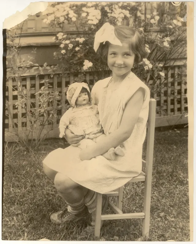 A sepia-toned photograph of a young light-skinned girl sitting outside in a chair, smiling and cradling a doll. She wears a light color dress with a large bow in her hair. Behind her is a lattice and flowering plants.