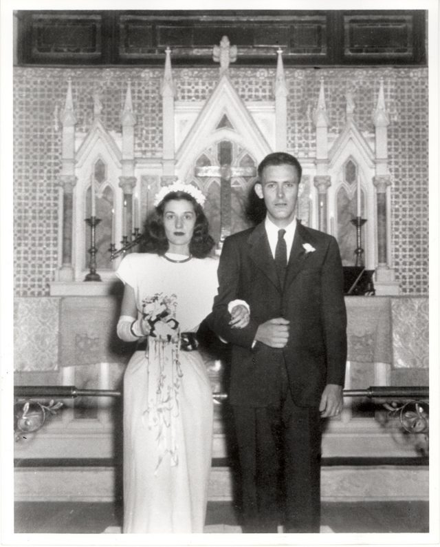 A black-and-white wedding portrait features a light-skinned, slim couple standing in front of an ornate altar. The woman clasps her husband, clad in a suit, at the elbow and holds a bouquet. Neither smile.