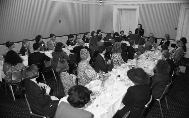 A black and white photograph of a room full of men and women seated at two long tables. In the front of the room a light-skinned woman with short brown hair stands at a podium addressing the group. The men are wearing suits and the women are wearing formal luncheon attire from the 1980s.