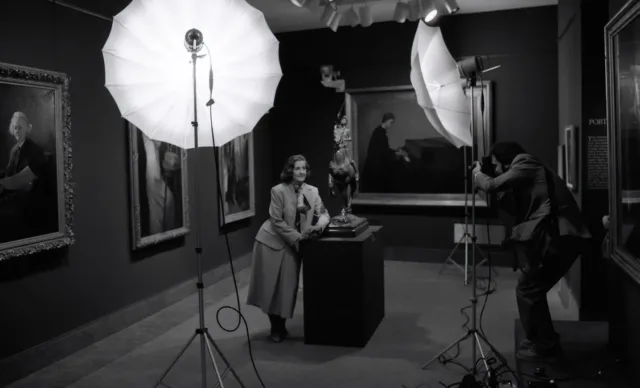 In an art gallery featuring five large oil paintings on the walls, a male photographer takes a posed photograph of a light-skinned woman leaning against a podium in the center of the room. It holds a sculpture of a man on a horse. The woman smiles slightly under the photographer's large umbrella lights. She wears a blazer, long skirt and an elegant silk scarf tied around her neck.