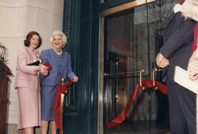 Two older light-skinned women in suit and skirt sets smile and cut a red ribbon on the doors of an institution. One woman has brown hair and the other hair white hair.