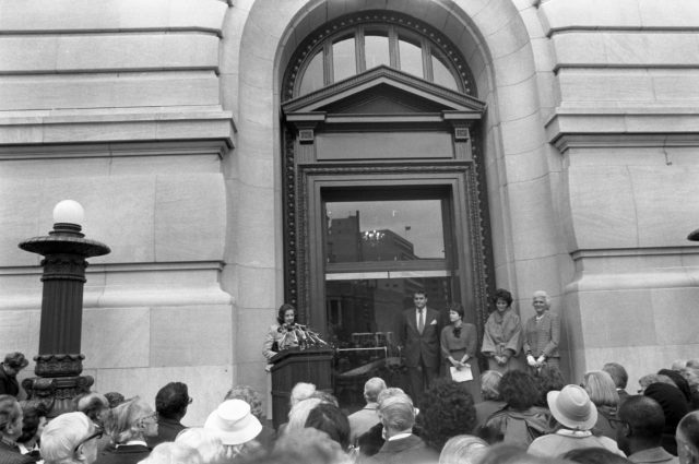 A light-skinned woman speaks at a podium adorned with many microphones outside of a large building. A crowd of people look on and four others, three women and a man, stand in front of the crowd, to the side of the woman with their hands clasped in front of them.