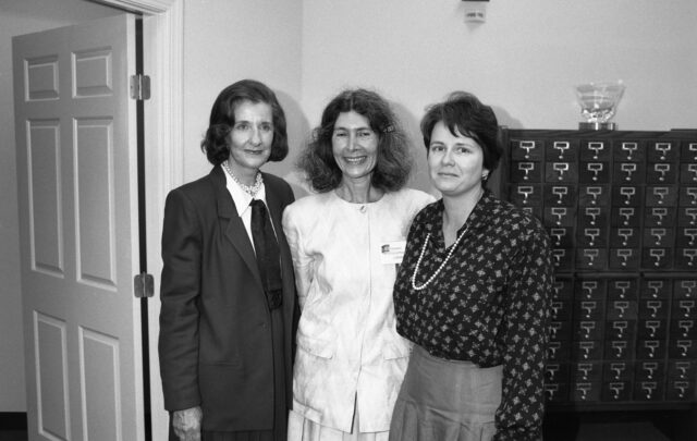 Three light-skinned women stand in a row smiling. They all have short to medium-length dark hair and wear 1980s-style business wear. Two wear pearls. In the background is a wooden library card catalogue.