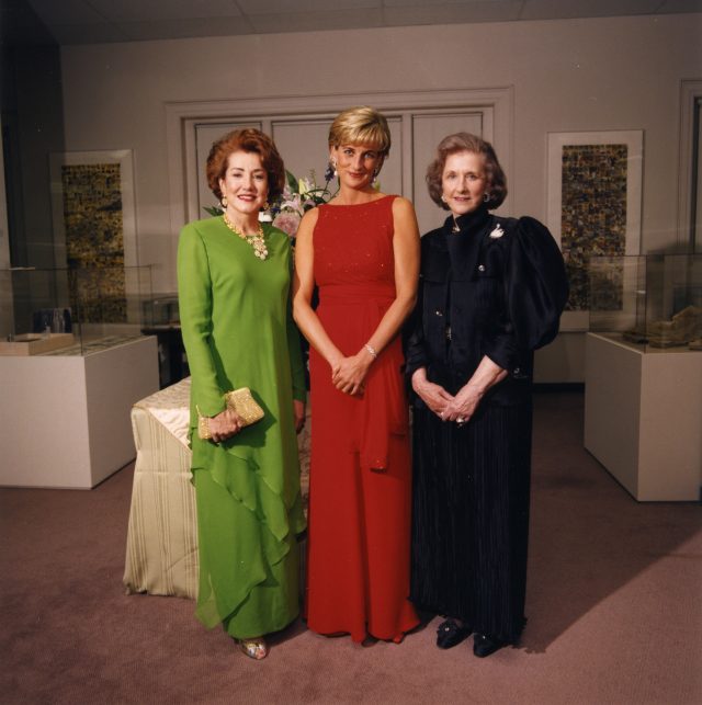 Three light-skinned women stand next to each other in fancy evening dresses inside an art gallery. The woman on the left wears a green dress, has red hair, and clasps a purse in her left hand; the women in the middle has short blonde hair, wears a red dress, and clasps her hands together in front of her; the women on the right has greying brown chin-length hair and wears a black dress, and also clasps her hands together in front of her. They all smile at the camera.