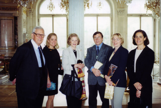 A color photograph of a group of 2 light-skinned men and 3 light-skinned women who stand in an ornate room, with large windows, crystal chandeliers, colored marble details, and gold accents. The figures are all wearing business attire and smiling at the camera.