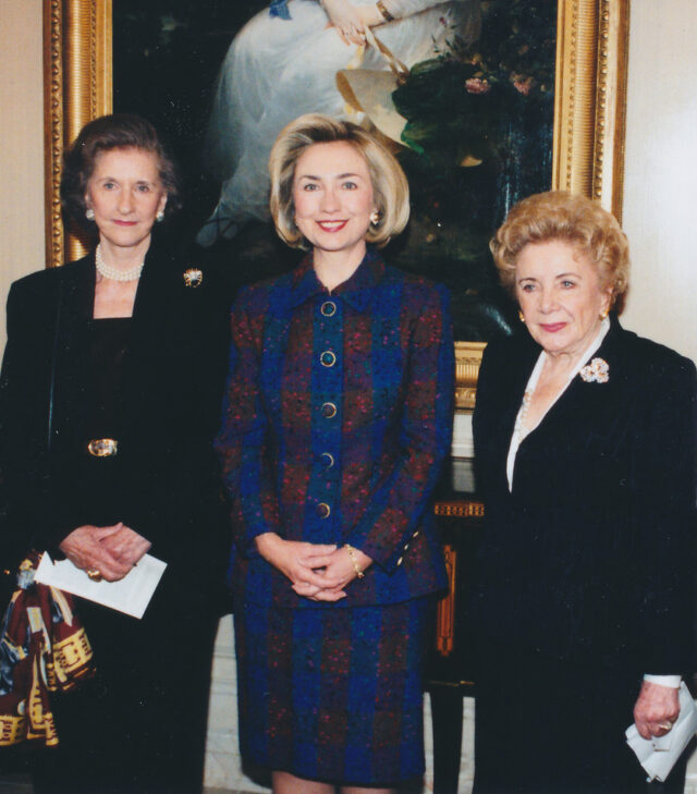 Three light-skinned women wearing formal skirt and blazer sets stand in a row smiling for a photograph. A woman with blonde hair wears a blue and red/orange striped suit, while the others are all in black with different broaches adorned on their left lapel