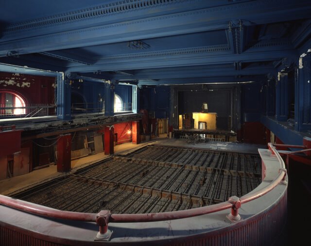 A large, ornate hall is photographed from a balcony, looking down onto a floor under renovation, with steel supports in place. Around the hall paint chips off of the walls and a layer of dust coats the balcony railing. Construction debris is visible on the left side.