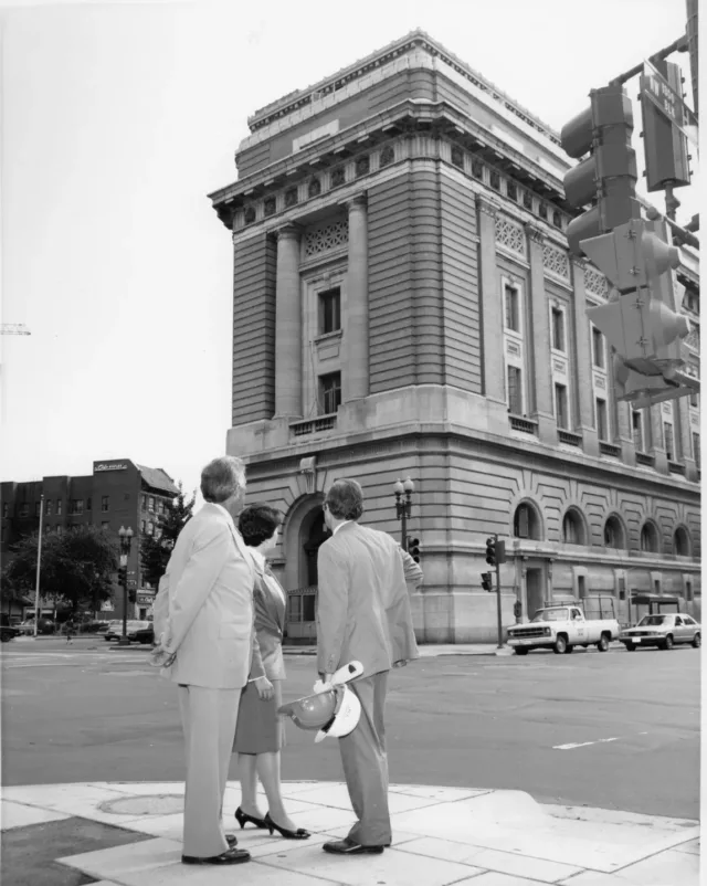 A black and white photograph of two light-skinned men and a light-skinned woman dressed in suits from the 1980s stand across the street from an ornate five-story stone building. The figures glance up a the building.