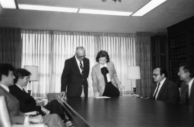 At the head of a large board table, a light-skinned woman stands next to a light-skinned man, both wearing business attire. Four other men sit at the table looking at the man and woman. The woman leans down slightly, signing a document with her right hand.