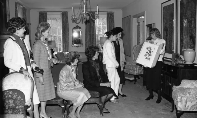 A group of light-skinned women in an ornate residential living room. One woman holds up a large book of illustrated prints and talks to the group. The other women look on with expressions of happiness and intrigue.