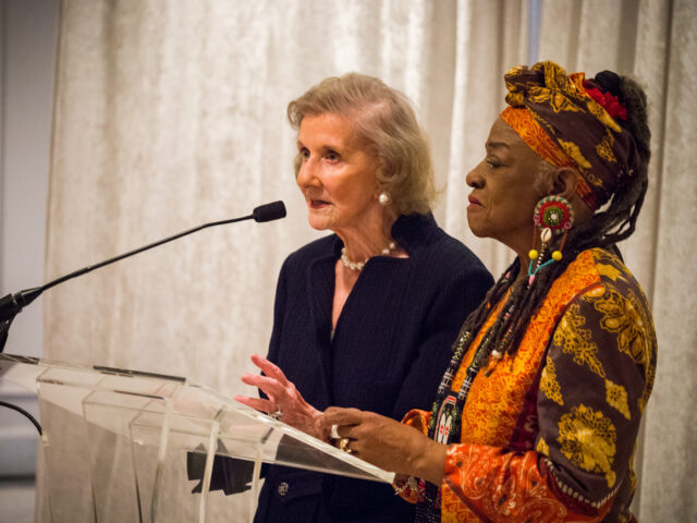 A light-skinned older woman speaks into a black microphone at a podium. Standing next to her is a black woman wearing a colorfully patterned matching headwrap and shirt and large beaded earrings.