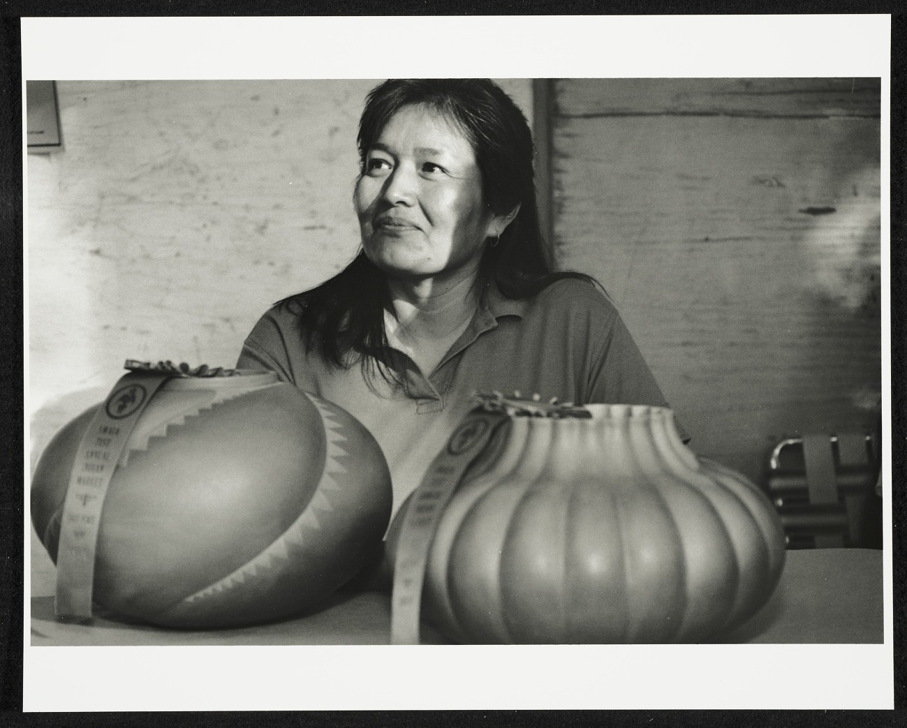 A black-and-white photo of a Native American woman with dark, long hair. She sits at a table with two large pieces of pottery in front of her, each has a winner ribbon on it. She looks off to the left smiling slightly as sunlight illuminates her face.