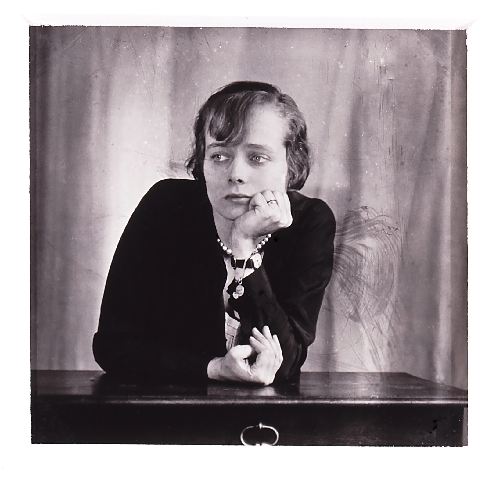 A black-and-white photograph of a light-skinned woman with short cropped hair leaning on a table with her left hand holding her chin. She looks off to the left, unsmiling.