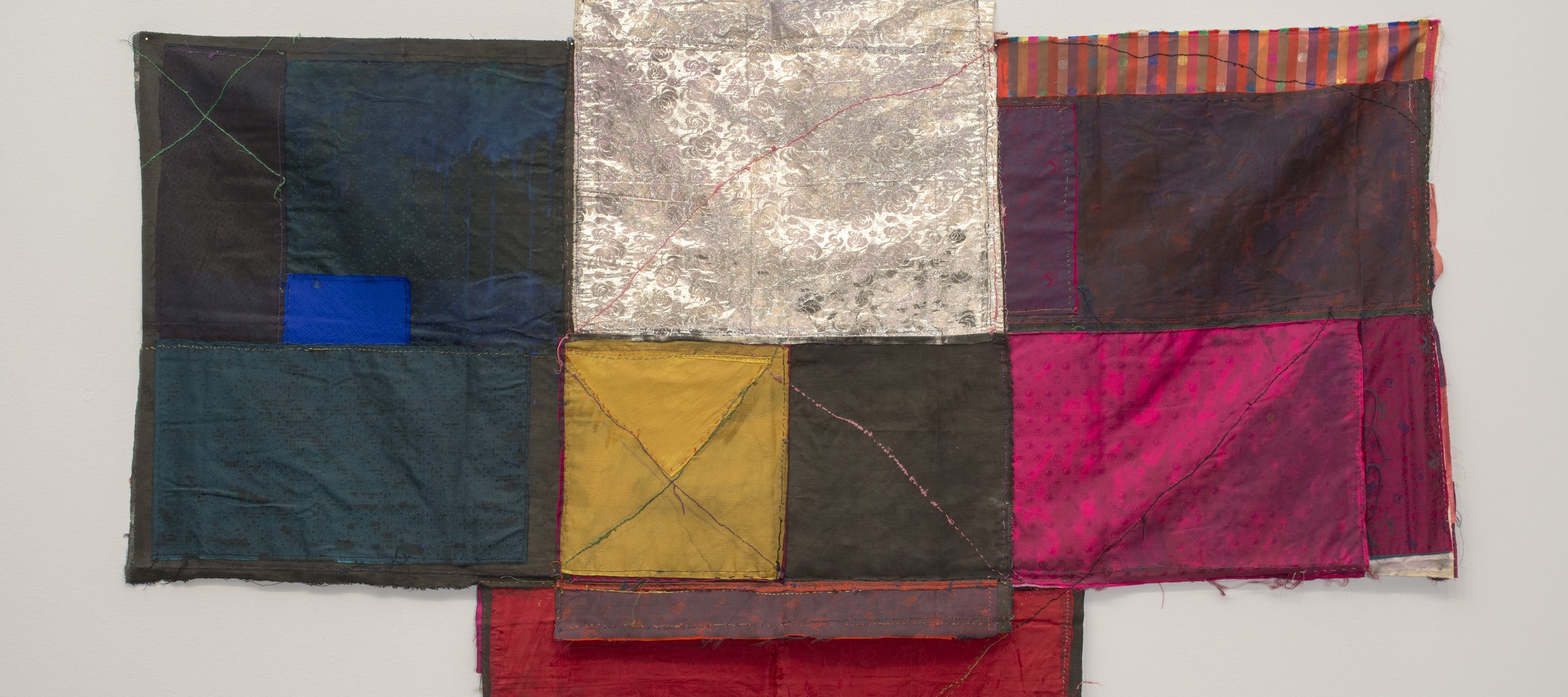 Textile collage of cloth folded and stitched together in loose geometric formations with visible seams making linear patterns. Deep blues on the left and warm colors on the right frame blocks of silver, yellow and grey in the center, which extends into a slightly longer base.