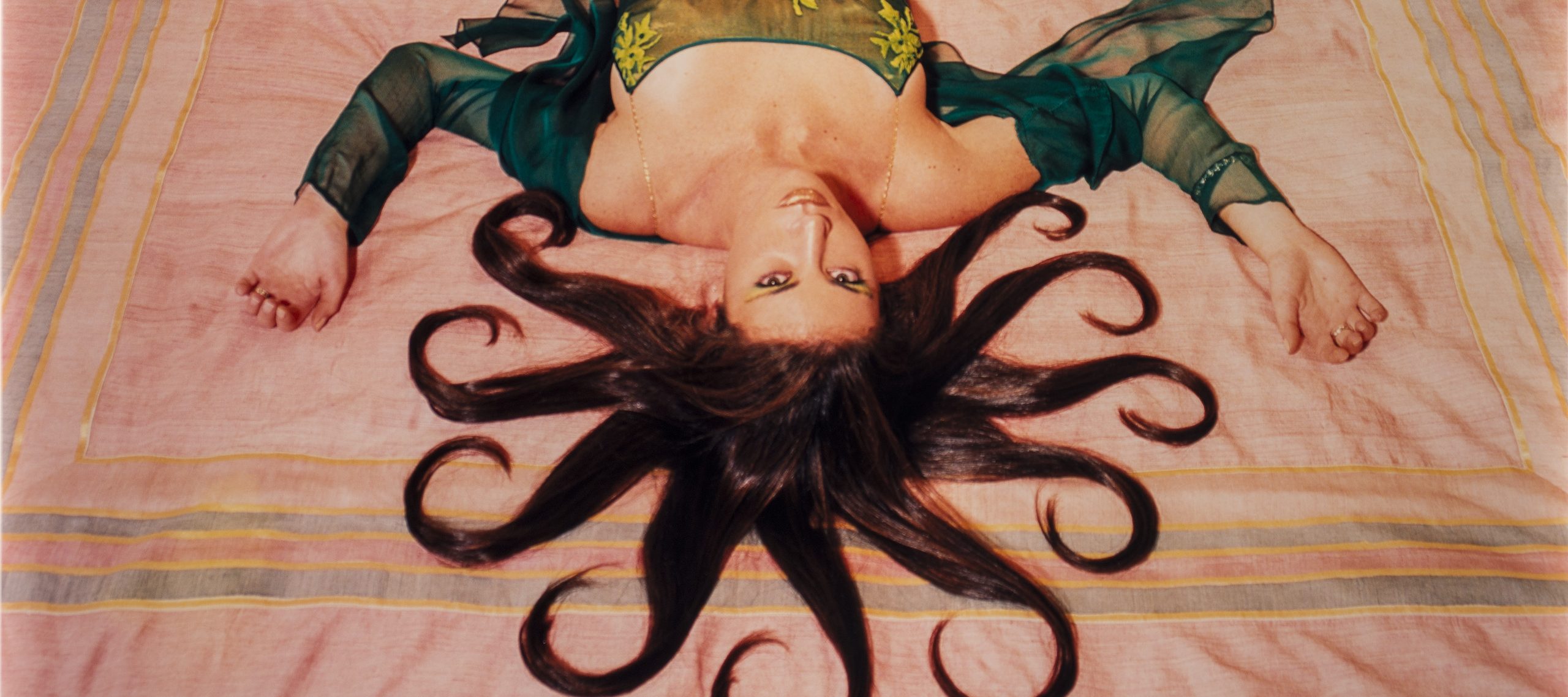 A slim, light-skinned woman wearing a long, emerald-green dress gazes up at the viewer from the foot of the bed, her bare feet near the headboard. Brown hair radiates from her head in 10 curled, snake-like segments. Her arms, bent at the elbows, extend from her body to form a W.