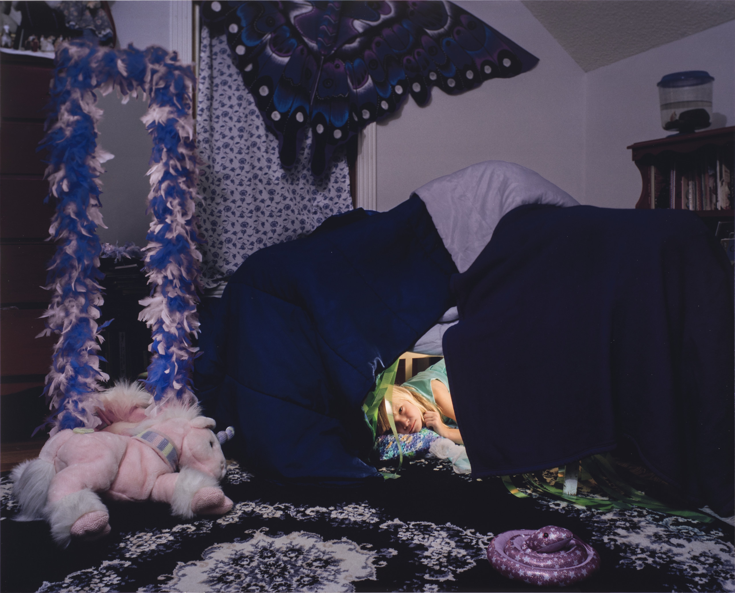 A color photograph of a young light skinned girl with light blonde hair. The girl peeks out from a fort made from a dark blue and purple comforter. A light pink and purple stuffed unicorn sits on a purple and white floral rug in front of the fort. A purple, blue, white, and black paper butterfly-shaped kite hangs on the wall above the fort.