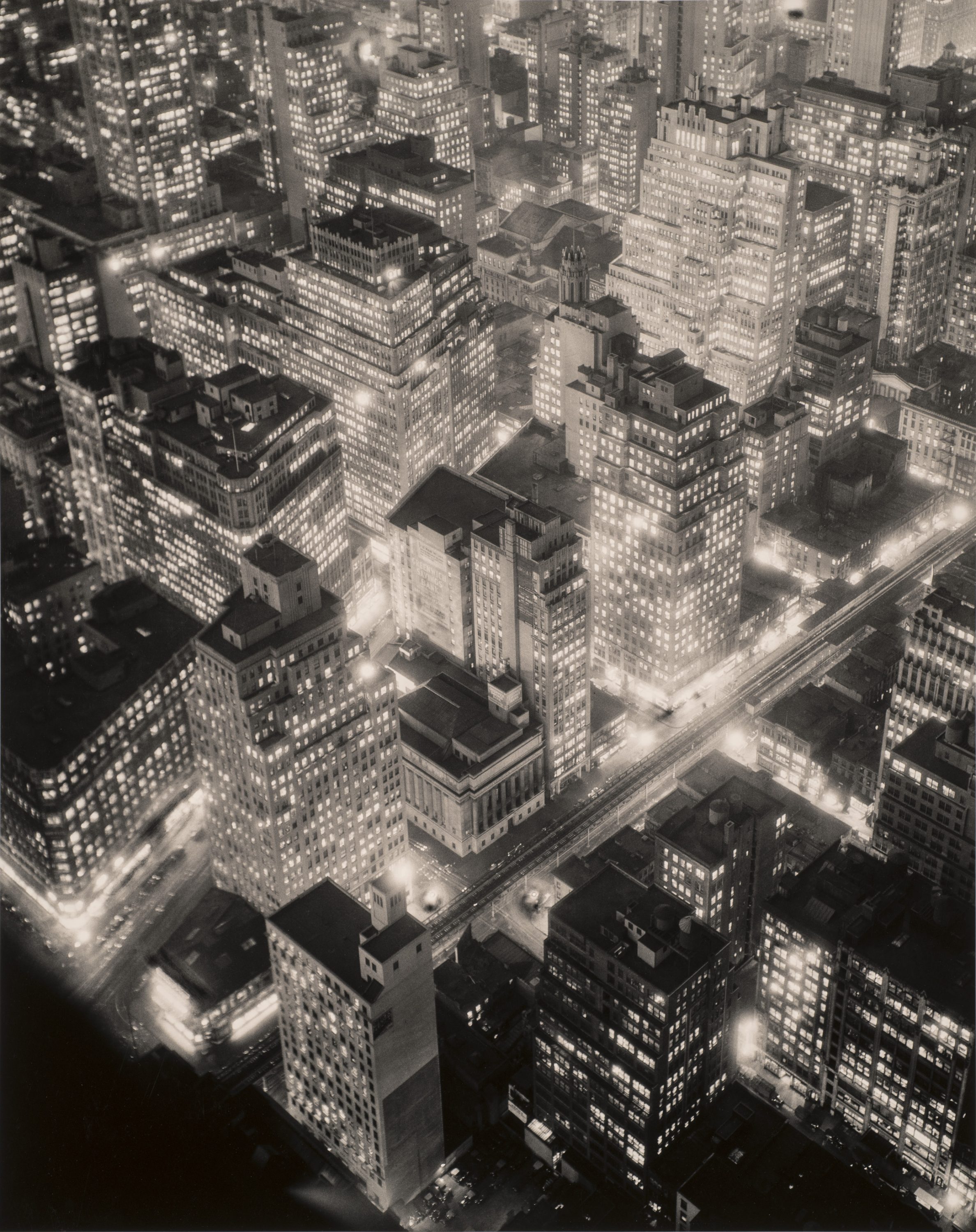 A black-and-white photograph of New York City seen from above at night. Towering, blocky skyscrapers emerge from the ground, glowing from tiny square windows of light, lining streets filled with blurs and flashes of light.