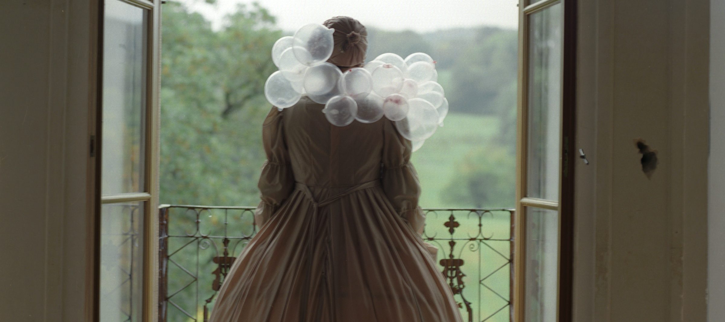 An adult woman seen from behind standing before two open glass doors leading out to a balcony overlooking greenery. She is dressed in a long, tan, period gown and her hair is tied up. Around her shoulders are several clear bubbles, resembling a scarf made of bubbles.