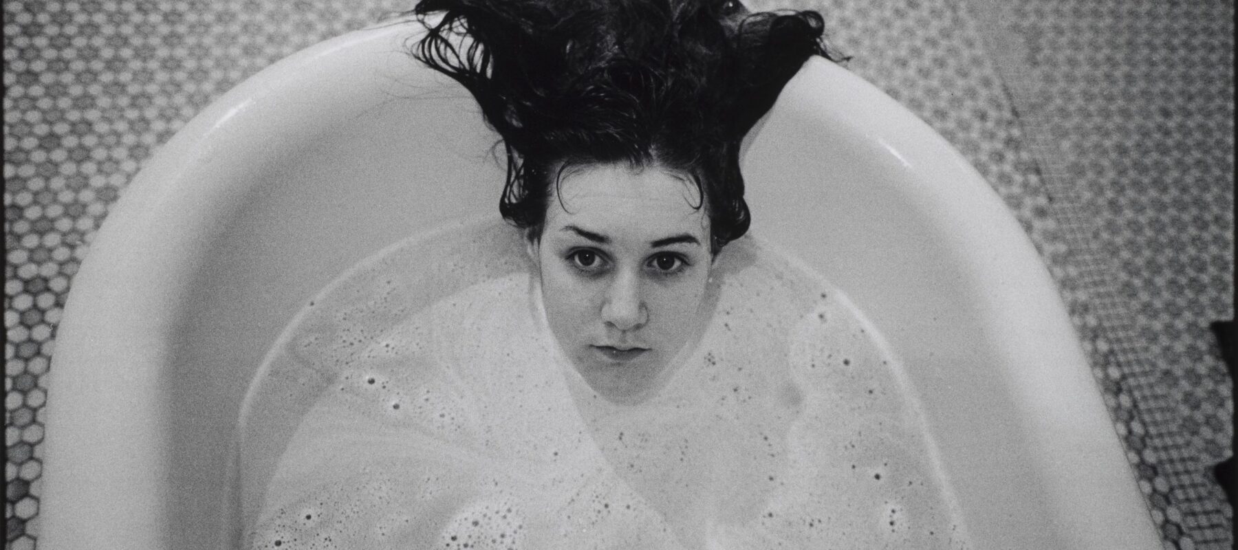 A black-and-white photograph of a light-skinned girl submerged in a white bathtub. Only her head is visible above the soap suds, and her dark hair hangs over the side of the tub. The floor beneath the tub is tiled.