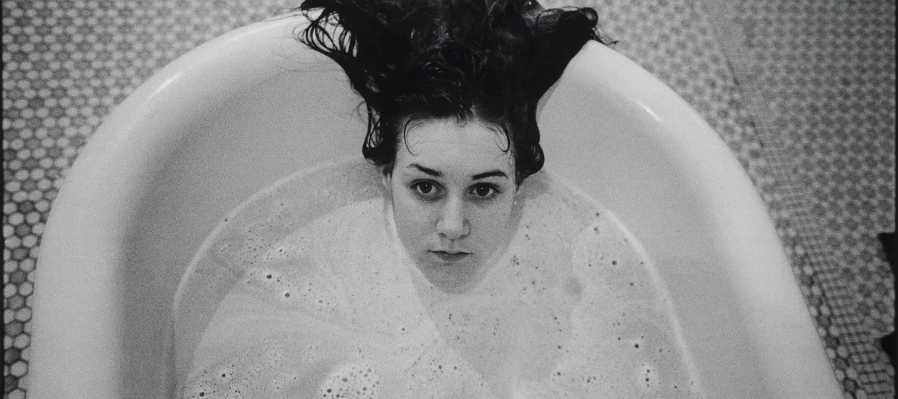 A black-and-white photograph of a light-skinned girl submerged in a white bathtub. Only her head is visible above the soap suds, and her dark hair hangs over the side of the tub. The floor beneath the tub is tiled.
