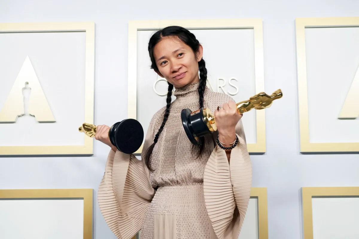 A medium-skinned woman of Asian descent holds two Oscar award statues in either hand, both pointed out to either side. She smiles slightly, her black hair is in two french braids, and she wears an earth-toned dress with fan sleeves.